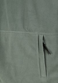 Close-up van grijze fleece stof met een verticale ritszak voorzien van een zwarte trekkoord.