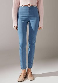 Högmidjade ljusblå jeans i denim med smal passform, subtil stretch och ofulländad fåll. Bärs med en rosa kortärmad tröja och beige skor.