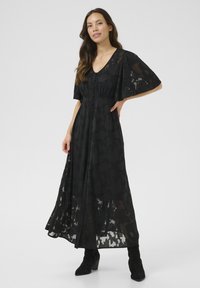 Abito maxi nero con maniche corte a forma di ala, motivo floreale, scollo a V e dettagli con bottoni che corre lungo la parte anteriore. Indossato con stivaletti neri alla caviglia.