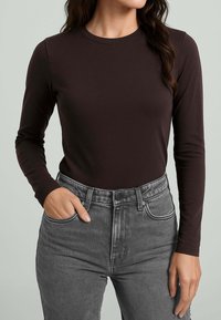 Top aderente a maniche lunghe in tessuto marrone scuro, abbinato a jeans grigi a vita alta. Scollo rotondo strutturato e dettagli di cucitura visibili.