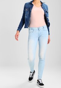 Jeans skinnies de color azul claro con textura desgastada, combinados con una blusa de cuello en V de color rosa claro y una chaqueta de mezclilla oscura. Zapatillas negras completan el conjunto.