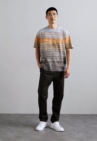 Striped short-sleeve t-shirt in gray, orange, and brown tones. Paired with black trousers and white sneakers. Model stands upright against a neutral background.