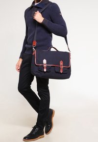 Granatowa torba na ramię wykonana z płótna, z brązowymi skórzanymi akcentami, metalowymi elementami i paskiem. Posiada zapięcia na klamry z przodu oraz zewnętrzne kieszenie.