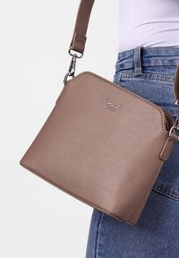 Křemově zbarvená texturovaná crossbody taška se stříbrným logem nošená přes rameno, v kombinaci s modrými denimovými džíny a bílým topem na jednobarevném pozadí.