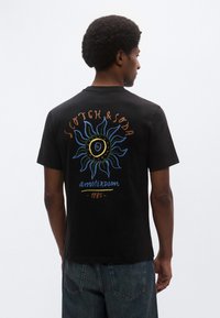 Μαύρο βαμβακερό t-shirt με ένα πολύχρωμο γραφικό ήλιου στην πλάτη και το κείμενο "Scotch & Soda Amsterdam 1985" με έντονα, φωτεινά γράμματα.