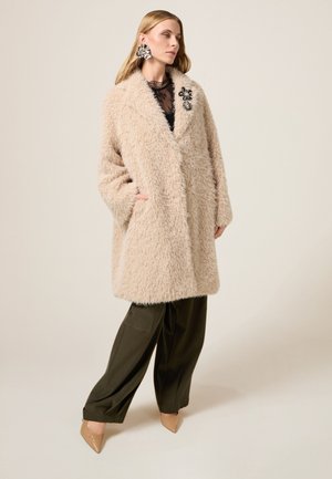 Beżowy puszysty płaszcz z ko�łnierzem, dużymi kieszeniami i ozdobną kwiatową broszką, w zestawieniu z luźnymi oliwkowo-zielonymi spodniami i beżowymi obcasami.