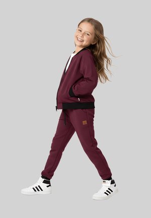 Młoda dziewczyna uśmiecha się podczas chodzenia, ubrana w bordowy dres i białe trampki z czarnymi paskami, na jednolitym szarym tle.