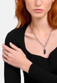 Silberne Halskette mit einem Anhänger aus schwarzem Kristall. Das Modell trägt ein schwarzes, geripptes Oberteil und einen silbernen Ring mit geometrischen Details.