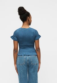 Μπλε τοπ άκι με σμόκιν, κοντά μανίκια με βολάν, υφή υφάσματος και εφαρμοστή μέση, συνδυασμένο με ψηλόμεσα τζιν παντελόνια σε ανοιχτό μπλε.