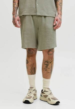 Pantalones cortos tejidos en verde oliva con un patrón texturizado, combinados con calcetines crema y zapatillas beige, con tatuajes prominentes en las piernas.