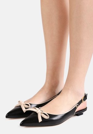 Pieds portant des ballerines slingback noires à bouts pointus avec des nœuds beiges et un détail de boucle dorée sur fond clair.