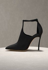Chaussure femme noire à talon haut avec bout pointu, bride à la cheville avec boucle et talon aiguille sur une surface claire avec un fond neutre.