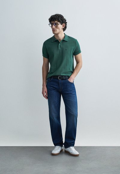 Polo vert foncé à manches courtes avec un col, porté avec un jean bleu et des baskets blanches. Le modèle est debout, posé contre un fond gris.