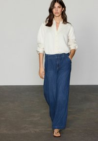 Hvid skjorte med oprullede ærmer, parret med løse, blå denim bukser med bred ben. Outfittet suppleres af enkle sandaler.