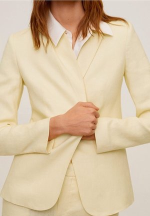 Femme portant un blazer jaune clair ajusté sur une chemise blanche à col, serrant le blazer à la taille.