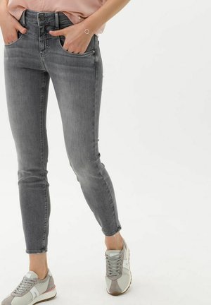 Kvinde iført grå skinny jeans, lyserød top og beige-grå sneakers med hænderne i lommerne, stående mod en hvid baggrund.