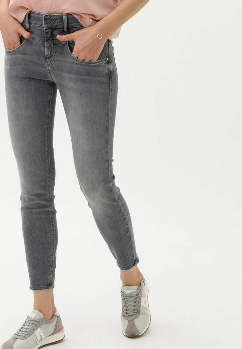 Femme portant un jean skinny gris, un top rose et des baskets beige-gris, les mains dans les poches, debout devant un fond blanc.