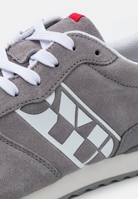 Gray suede sneaker with white laces, white logo accents, and a red heel tab. Features a textured surface and a lightweight sole design.