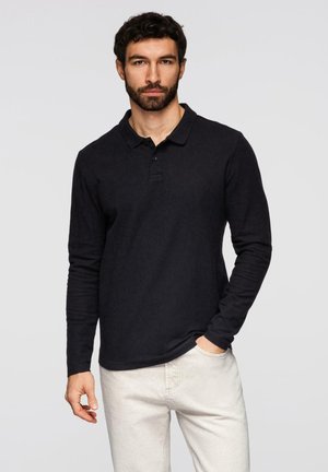 Mann mit dunklem Haar und Bart, trägt ein schwarzes Polohemd mit langen Ärmeln und helle Hosen, steht vor einem schlichten hellen Hintergrund.