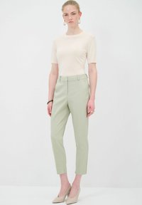 Béžový top s krátkým rukávem v kombinaci se světlými zelenými kalhotami na míru. Kalhoty mají slim fit střih a záhyb vpředu. Modelka má na sobě nahé vysoké podpatky.