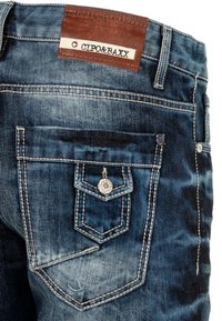 Jean en denim bleu foncé avec une poche arrière de forme unique ornée d'un bouton en argent, des coutures contrastantes et une étiquette en cuir sur la ceinture.