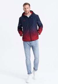 Capuchonjack in een marineblauwe en rode gradient. Heeft elastische manchetten en een ritsluiting. Draagt over een wit shirt met lichtblauwe spijkerbroek en witte sneakers.