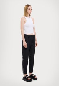 Weißes ärmelloses Crop-Top, schwarze tapered Hose mit elastischem Bund und schwarze Sandalen mit doppeltem Schnallen-Detail und strukturierten Sohlen.