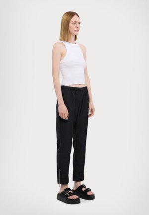 Hvit ermeløs crop topp, svarte smale bukser med elastisk midje, og svarte sandaler med dobbel spenne-detalj og teksturerte såler.