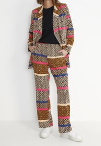 Vzorovaný oblek vyrobený z lehké látky; obsahuje geometrické tvary v béžové, hnědé, růžové a modré; zahrnuje černý top a bílé tenisky.