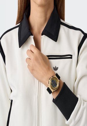 Oseba v beli jakni z črnim ovratnikom in manšetami, ki prilagaja ovratnik z roko, na kateri se sveti zlata digitalna zapestnica s črnim trakom.