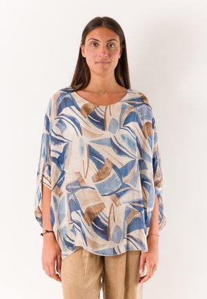 Donna con capelli lunghi e scuri indossa pantaloni beige e una maglia larga con motivi astratti a pennellate blu e marroni, in piedi su sfondo bianco.