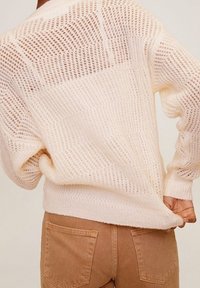 Pull en tricot beige clair avec un motif à maille ouverte, doté d'un large décolleté et de manches longues, associé à un pantalon texturé de couleur tan.