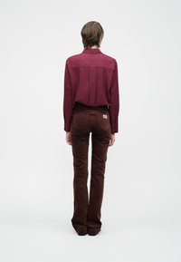 Bordová košeľa s dlhým rukávom a hnedé kárované nohavice s rozšírenými nohavicami, zobrazené zozadu. Nohavice majú zadné vrecká a štítok na pravej strane.