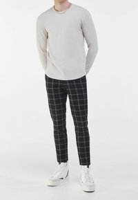 Světlé béžové tričko s dlouhým rukávem, přiléhavé černé kostkované kalhoty a bílé kotníkové tenisky. Minimalistický design se stříbrným řetízkem na krk.