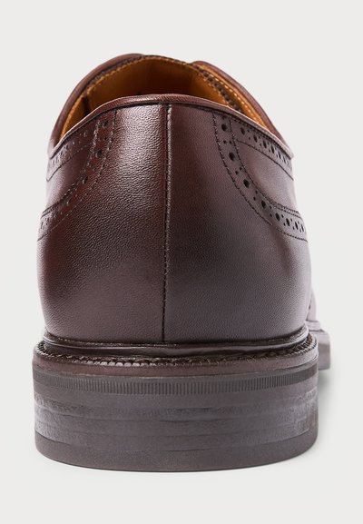 Smeđi kožni cipele s potpeticom i brogue detaljima, šivana potplata i stavljena drvena potpetica na bijeloj pozadini.