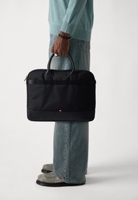 Schwarze Laptoptasche mit zwei Griffen, aus Stoff mit glatter Textur. Verfügt über einen Farbakzent am Boden und eine minimalistische Markenkennzeichnung.