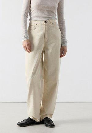 Inimene, kes kannab kõrge vöökohaga lahtisi koorevärvi pükse, helesinist pikkade varrukatega pluusi ja musti loafersi, seisab valge tausta ees.