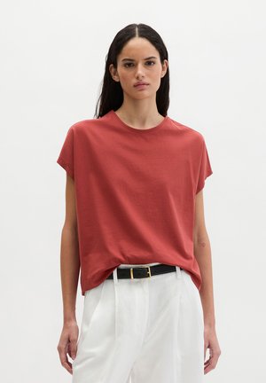 Jonge vrouw met lang donker haar draagt een losse rode top met korte mouwen, in een broek met hoge taille en witte kleur, afgewerkt met een zwarte riem.