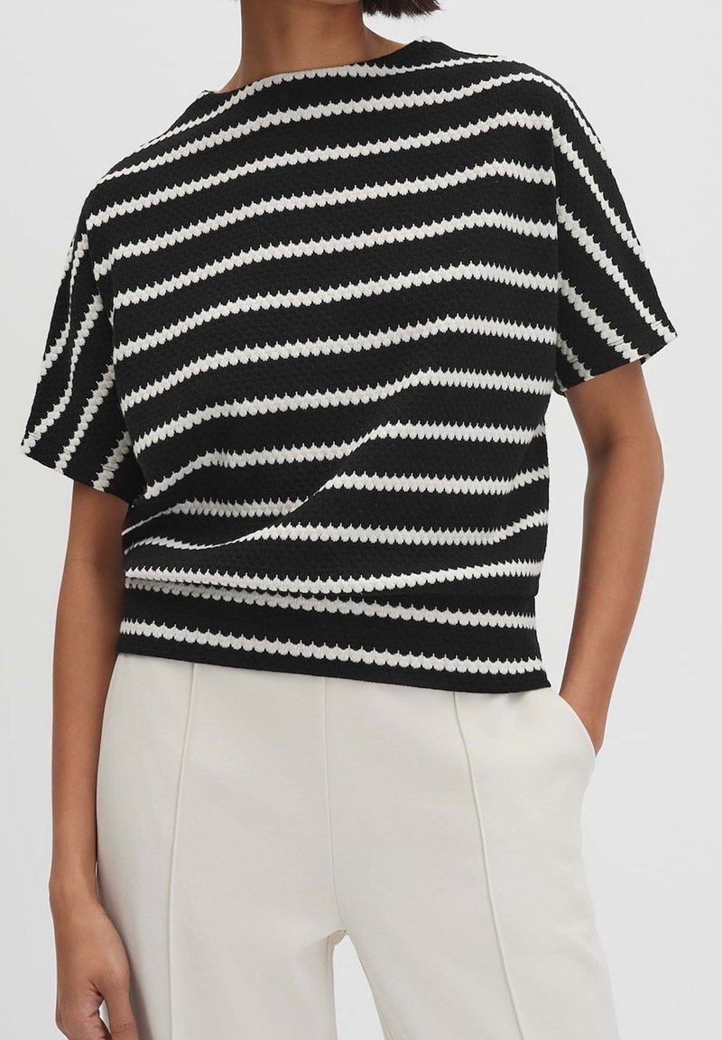 Personne portant un haut en tricot à rayures horizontales noires et blanches à manches courtes, associé à un pantalon blanc taille haute, une main dans la poche.