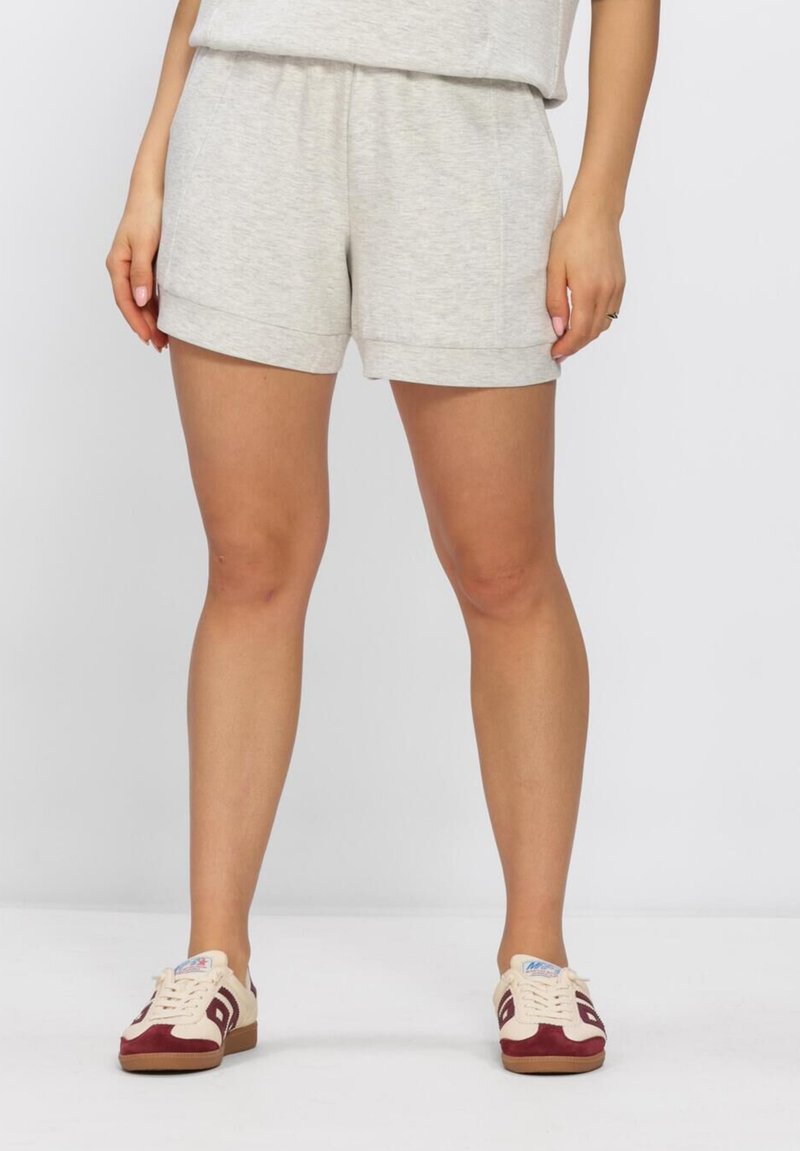 Woman wearing light gray casual shorts and beige sneakers with maroon accents, standing against a plain white background.