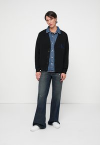 Black cardigan with buttons over a light blue denim shirt, paired with wide-leg faded blue jeans and white sneakers. Simple branding detail visible.