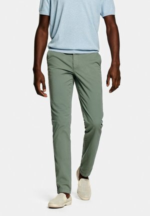 Pantalon chino en coton vert avec une coupe droite, doté d'une taille classique et d'une braguette zippée, assorti à des chaussures beige à enfiler.