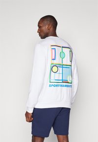 Sweatshirt blanc avec un motif géométrique coloré au dos, portant le texte "SPORTIVAMENTE". Associé à un short bleu marine.
