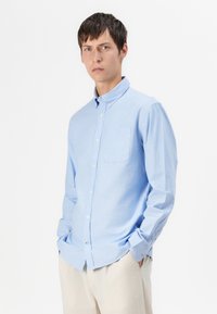 Camisa abotonada azul claro hecha de algodón suave, con cuello puntiagudo, un bolsillo en el pecho y mangas largas con puños abotonados.