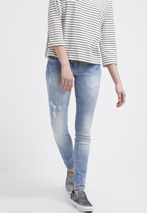 Personne portant un jean skinny déchiré bleu clair, un t-shirt à manches longues rayé noir et blanc et des baskets grises sans lacets marchant vers l'avant.