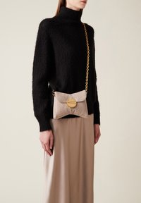 Beige Handtasche mit strukturiertem Material, einem runden goldenen Akzent und einer Kettenriemen.getragen zu einem schwarzen, flauschigen Rollkragenpullover und einem beigen Rock.