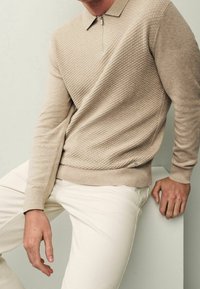 Beige pitkähihaisen poolopaidan yläosassa on tekstuuri ja vetoketjullinen kaulus. Paidassa on ribbineuloshelma, ja se on yhdistetty kermaisiin housuihin.