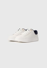 Fehér, alacsony szárú sneaker, sötétkék belső gallérral, fehér fűzőkkel és az oldalán található "Tommy Hilfiger" felirattal, egy sima fehér háttéren kiemelve.