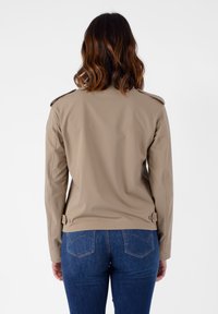 Giacca aderente beige con spalline, maniche lunghe e dettagli con bottoni in vita. Texture liscia, design minimal.