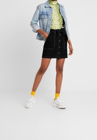 Žena v světle modré denimové bundě, zeleném vzorovaném roláku, černé sukně s knoflíky a mašlí, žlutých ponožkách a bílých teniskách.